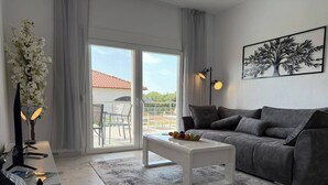 Un salon moderne avec un canapé gris, une table basse blanche et une grande fenêtre donnant sur un bâtiment au toit rouge.