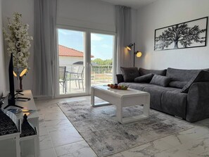 A modern living room with a grey sofa, a white coffee table, and a large window with a view of a red-roofed building. - Asteri Villas - 3 Bedrooms (Nea Propontida)