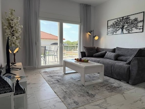 A modern living room with a grey sofa, a white coffee table, and a large window with a view of a red-roofed building. - Asteri Villas - 2 Bedrooms (Nea Propontida)