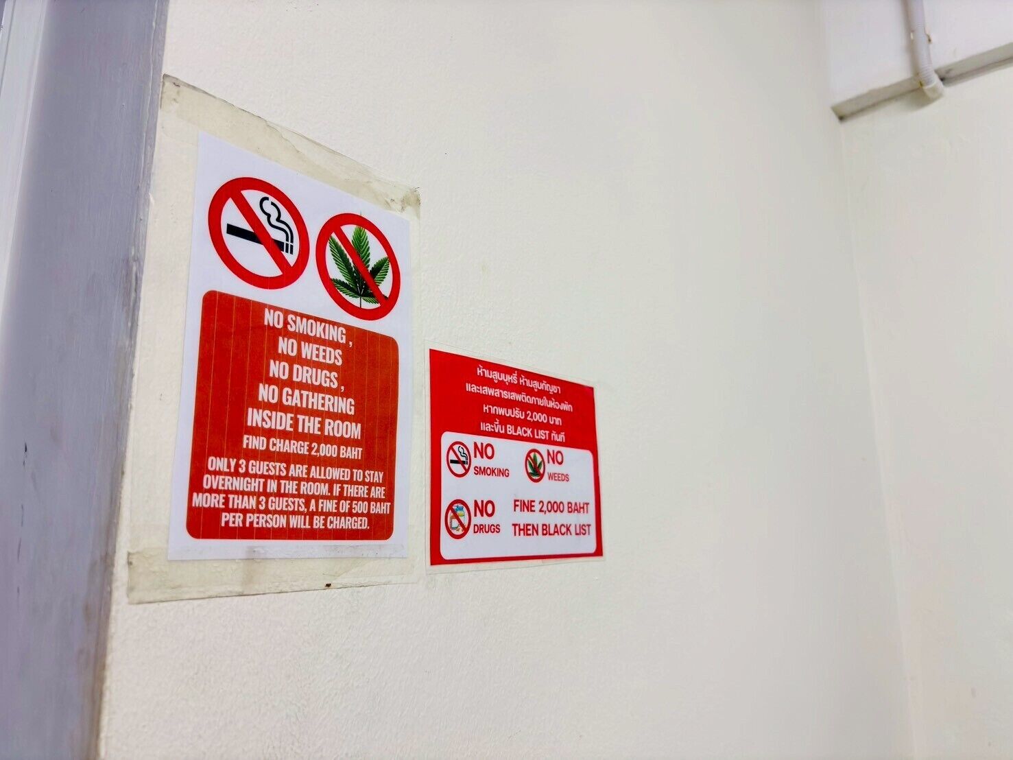 Dos carteles en una pared que indican prohibido fumar, prohibido el consumo de drogas, prohibido reunirse, y una multa de 2.000 baht por infracciones.