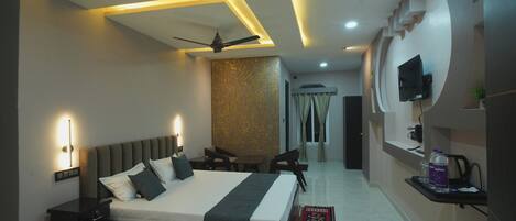 A modern hotel room with a large bed, bedside tables, a ceiling fan, a TV mounted on the wall, and a small seating area.
