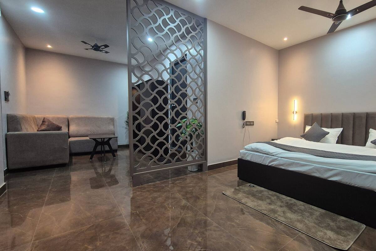 Un dormitorio moderno con cama, sofá, separador decorativo, ventilador de techo y luz empotrada en la pared.