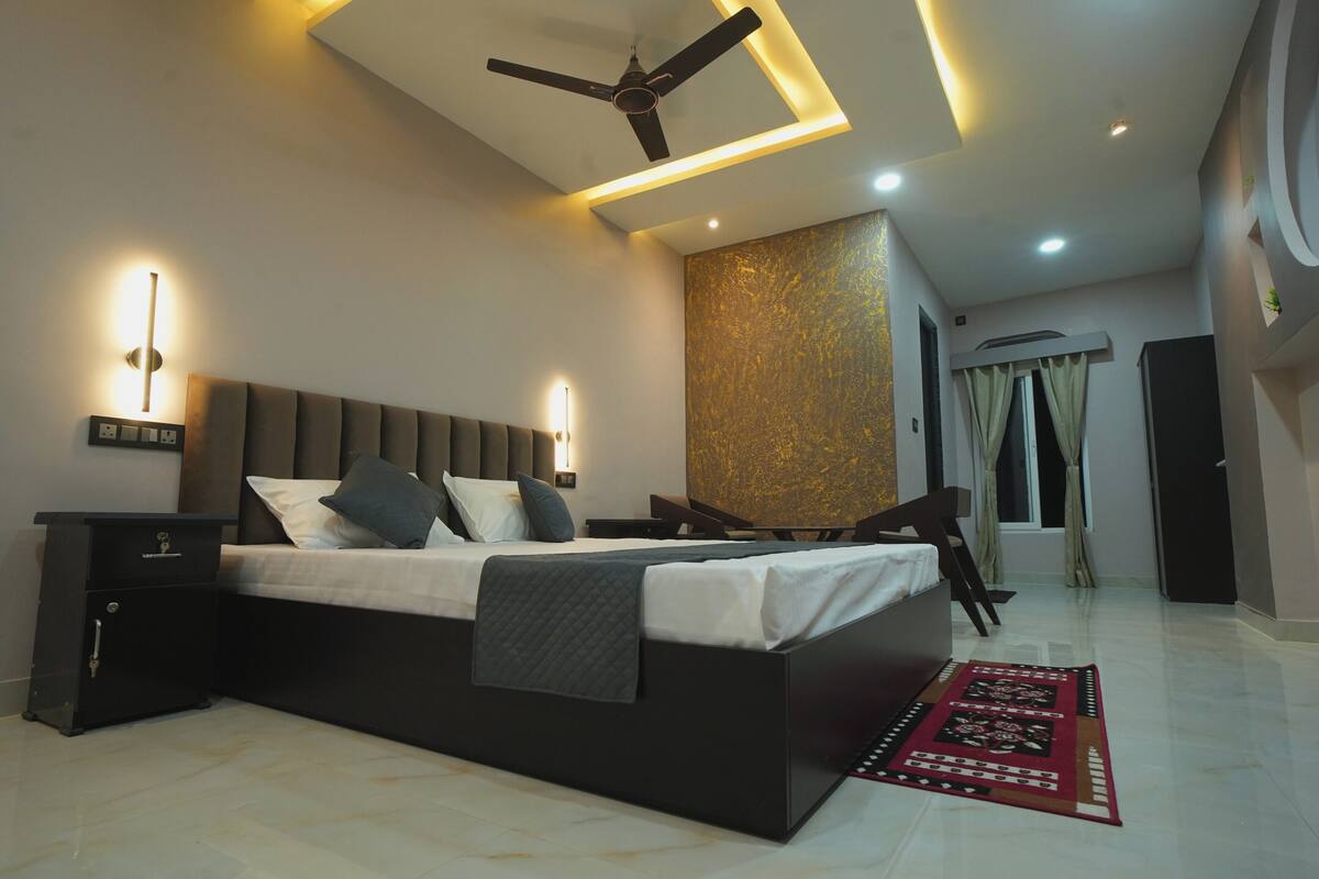 Un dormitorio moderno con cama, mesitas de noche, ventilador de techo, iluminación empotrada, un muro decorativo y una ventana con cortinas.