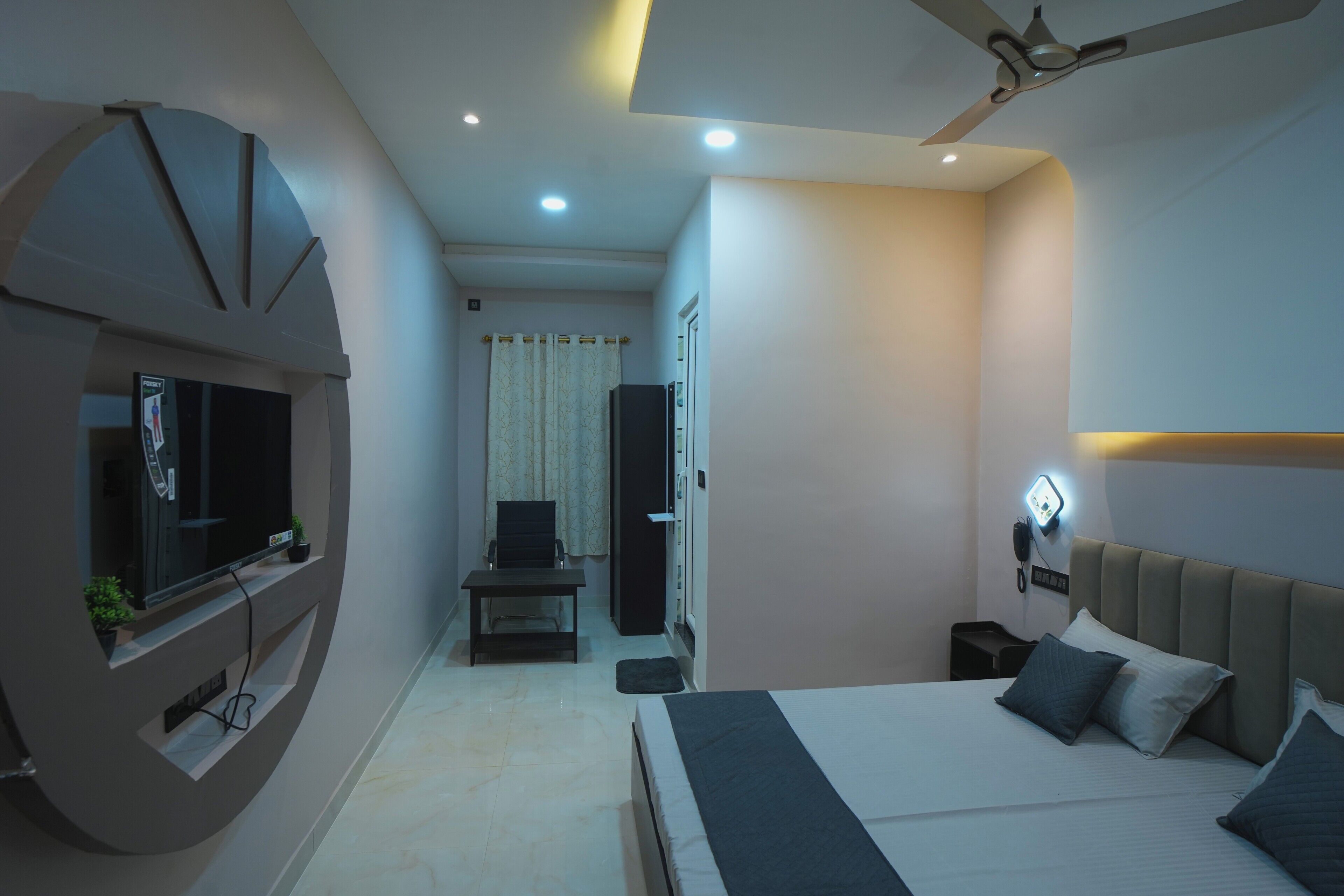 A modern hotel room with a bed, a TV mounted on the wall, a ceiling fan, and a bedside table.