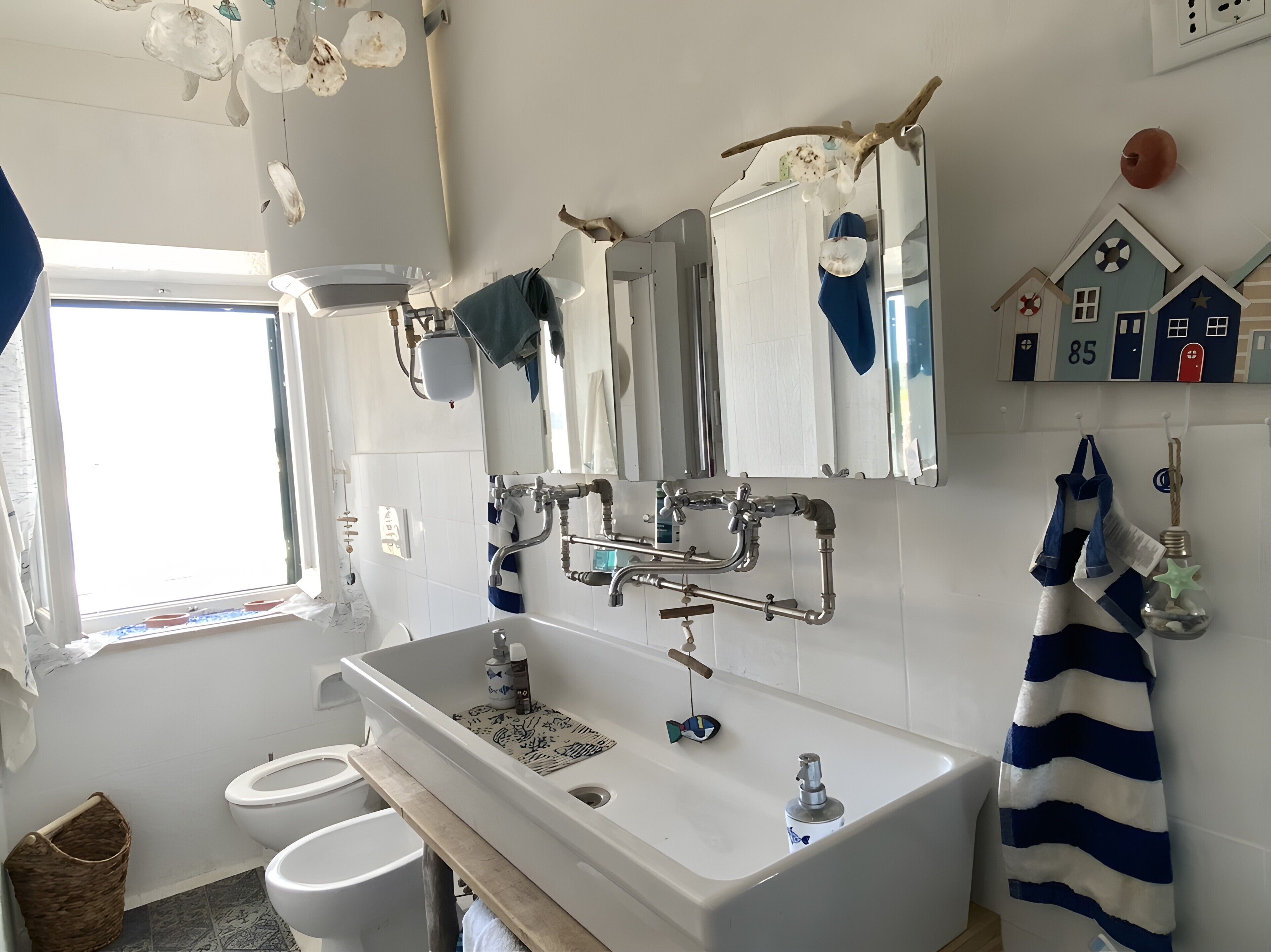 Un baño con un lavabo blanco, dos inodoros, una ventana y un espejo con una toalla colgada.