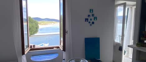 A room with a large window, a bed, a table with a bottle, a fan, and a view of the sea.