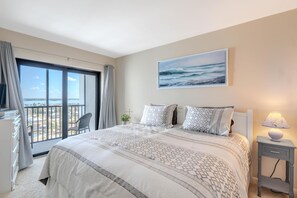 A bedroom with a large bed, a view of the ocean, a painting on the wall, and a nightstand with a lamp. - Island Winds Penthouse 1 (Fort Myers Beach)