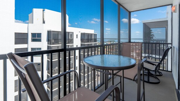 Een balkon met een glazen tafel en stoelen, met uitzicht op een stadsgezicht en de zee.