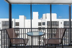 A balcony with a glass table and two chairs, overlooking a cityscape with white buildings. - Island Winds Penthouse 1 (Fort Myers Beach)