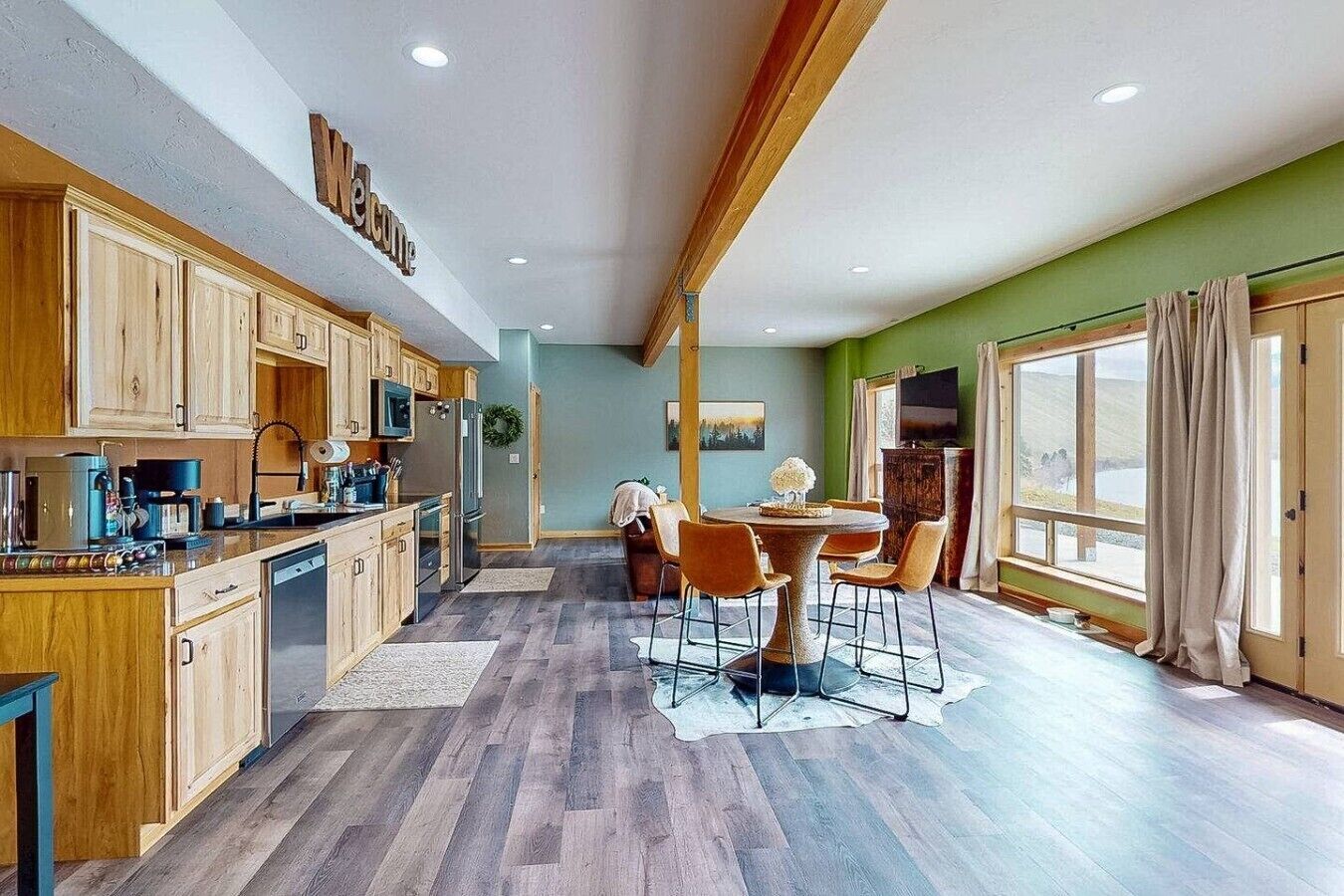 A spacious kitchen with wooden cabinets, a dining area with a round table and chairs, and a large window with curtains.