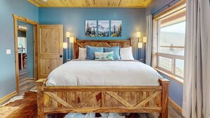 Een slaapkamer met een houten bedframe, een groot raam met gordijnen, een houten plafond en een deur naar een andere kamer.