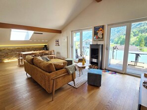 Ein großzügiger Wohnbereich mit einer Couch, einem Esstisch und einem Kamin. Große Fenster bieten Blick auf einen See und Berge.