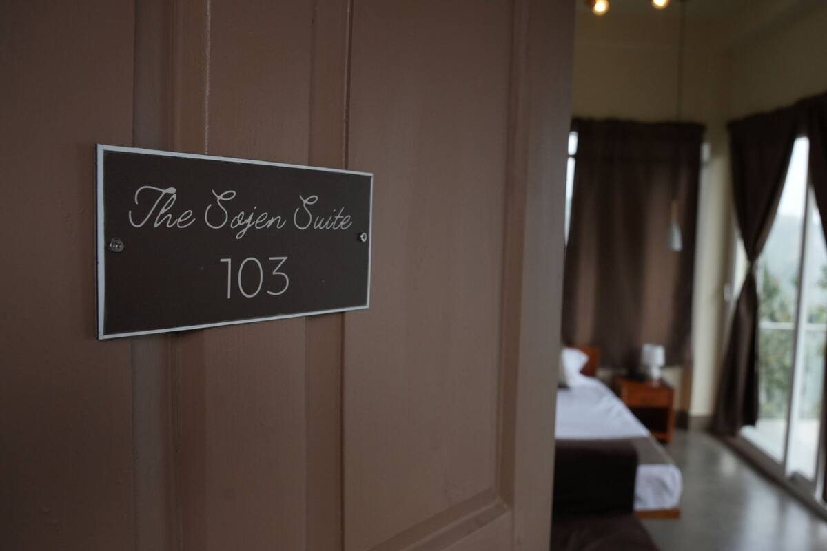 A sign indicating 'The Sojen Suite 103' in a hotel room with a bed, curtains, and a window.