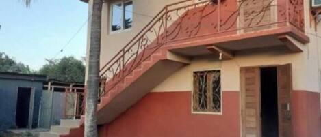 Un bâtiment à deux étages, avec une façade rouge et beige, un escalier menant à l’étage supérieur et une petite porte au rez-de-chaussée.
