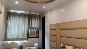 A room with a ceiling fan, a window with curtains, an air conditioning unit, a sofa, and a bed with a headboard. - Hotel Amrit Villa Karol Bagh New Delhi (New Delhi)