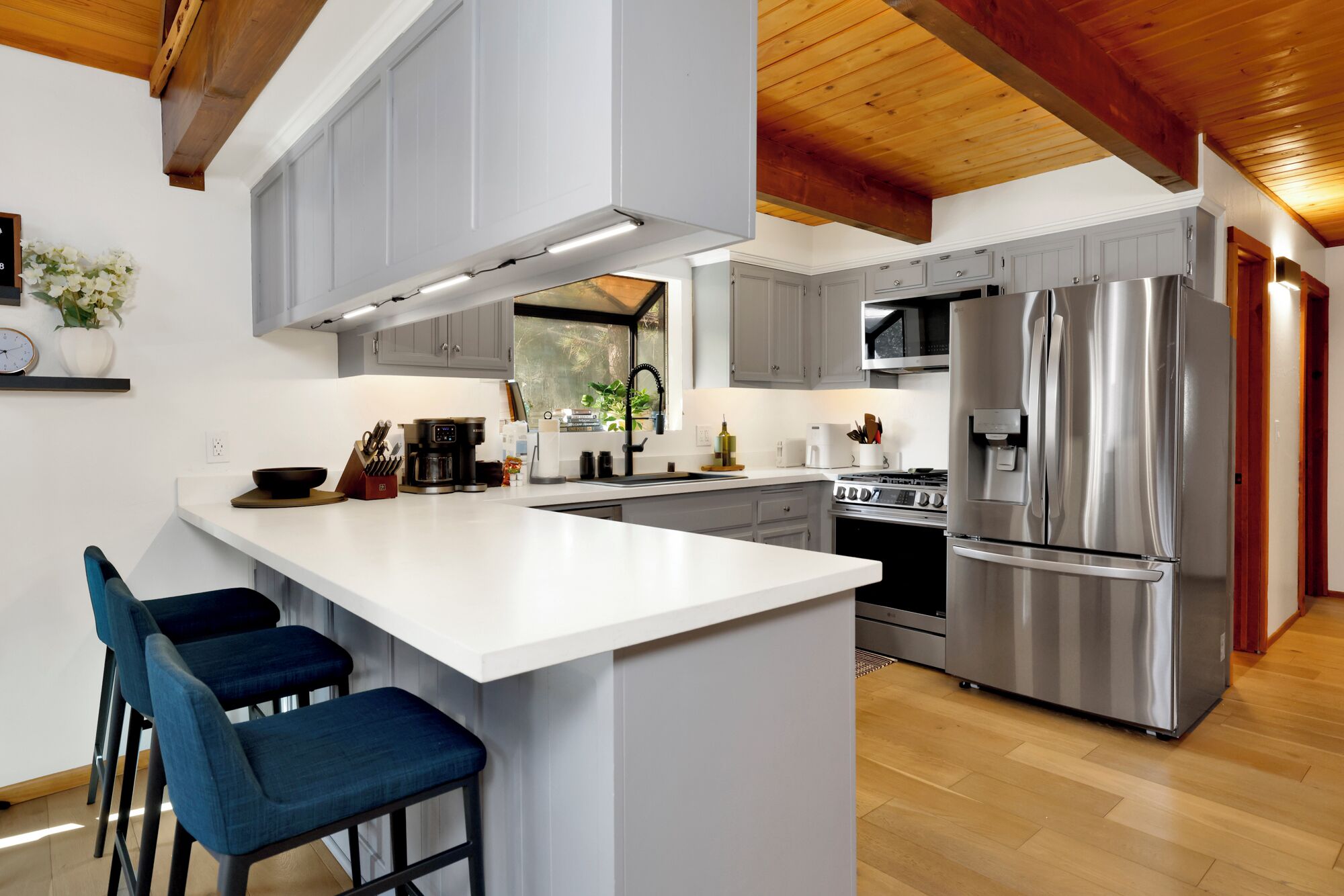 A modern kitchen with a central island, bar stools, stainless steel appliances, and a wooden ceiling.