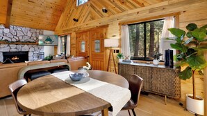 A wooden cabin interior with a stone fireplace, a dining area, and a kitchen. - Hot Tub Cabin w/ Game Loft, Fireplace + Deck – Near Lake & Slopes (Big Bear Lake)