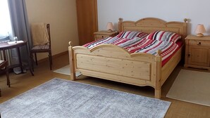 A bedroom with a wooden bed, two bedside tables with lamps, a chair, a desk, a window with curtains, and a door. - Cozy vacation apartment "Haus Bertram" in Leun. in a quiet rural location. (Leun)
