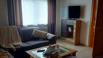 Olohuoneessa on sohva, kirjoja sisältävä sohvapöytä, jalustalla oleva televisio ja ovi toiseen huoneeseen.