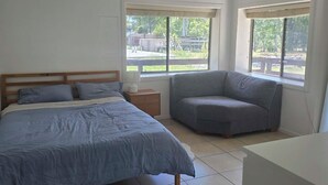Un dormitorio con una cama, una mesita de noche, un sillón gris y dos ventanas.