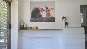 Una habitación con suelo de baldosas, una pared blanca, un cuadro enmarcado con dos perros, una estantería con objetos pequeños y un armario blanco.