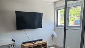 Habitación con televisión de pantalla plana montada en la pared, un mueble de televisión de madera con controles remotos, un escritorio blanco con una silla, un ventilador de pie y un ventanal corredizo que da al exterior.