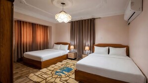 Habitación de hotel con dos camas individuales, cabecera de madera, alfombra estampada, lámpara colgante y unidad de aire acondicionado.