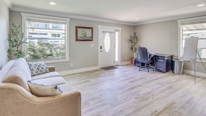A spacious room with a light-colored wooden floor, a beige sofa with patterned cushions, a desk with a chair, and a whiteboard. - Steps to Bayshore Blvd! Snowbird Retreat w/ Pool (Tampa)