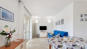 Een moderne woonkamer met een bank, een eettafel met een tafelkleed met patroon, een televisie en een vaas met bloemen op een houten kast.
