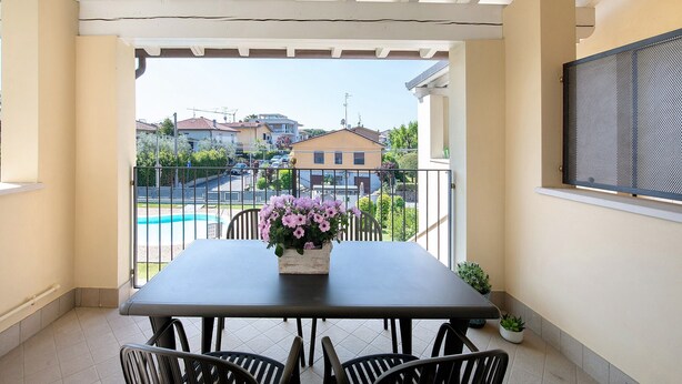Een balkon met een tafel en stoelen, een bloevas, een bloempot en uitzicht op huizen en een zwembad.