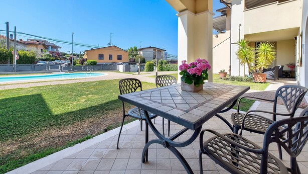 Un patio exterior con una mesa y sillas, un pequeño jardín y una piscina al fondo.