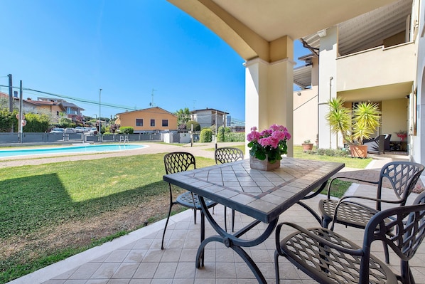 An outdoor patio with a table and chairs, a small garden, and a pool in the background.