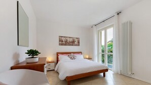 Un dormitorio con cama, mesitas de noche, una planta en maceta, una silla, ventana con cortinas y un radiador.
