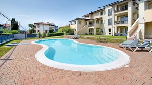Una piscina con solárium de ladrillo, sillas de descanso y un edificio de varios pisos con balcones.