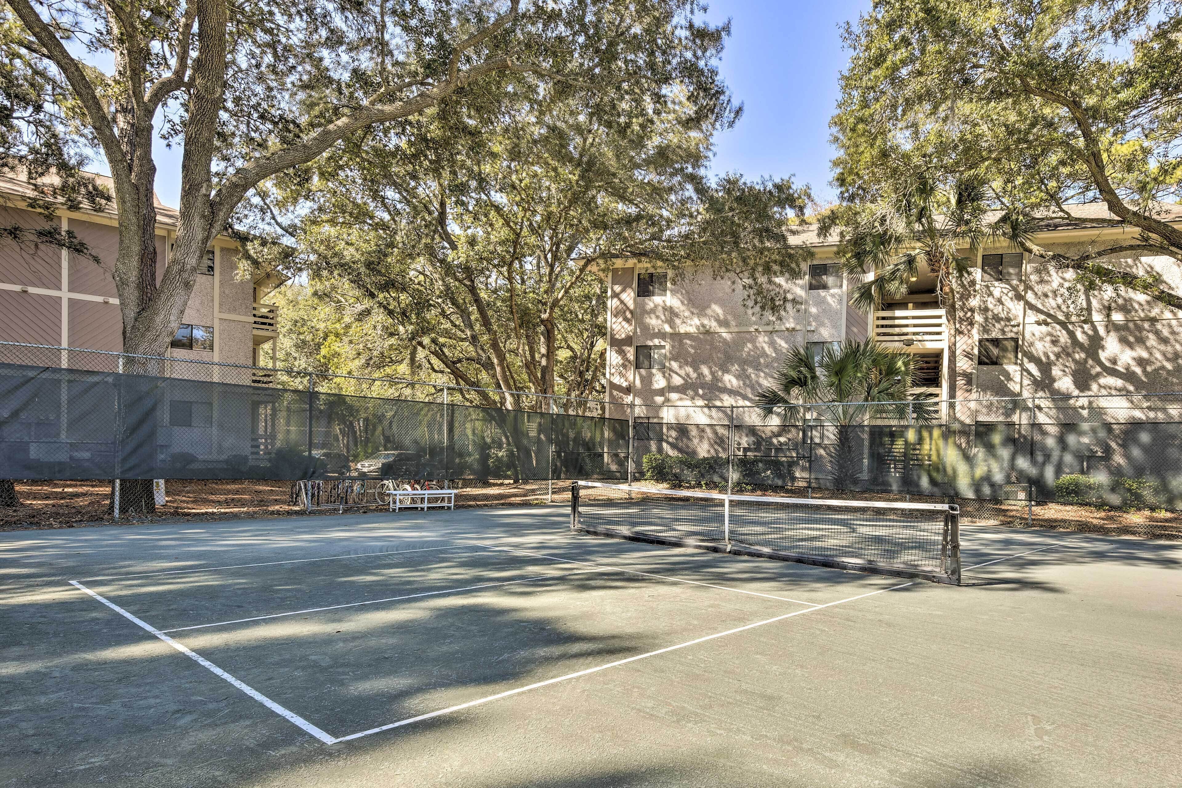 An outdoor tennis court with a net, surrounded by trees and a building in the background.