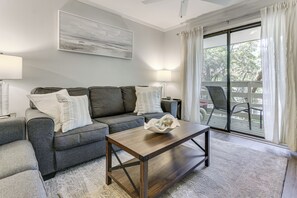 A living room with a sofa, coffee table, and a view through a sliding glass door. - 'folly Tides' HHI Condo w/ Beach, Pools & Tennis! (Hilton Head Island)