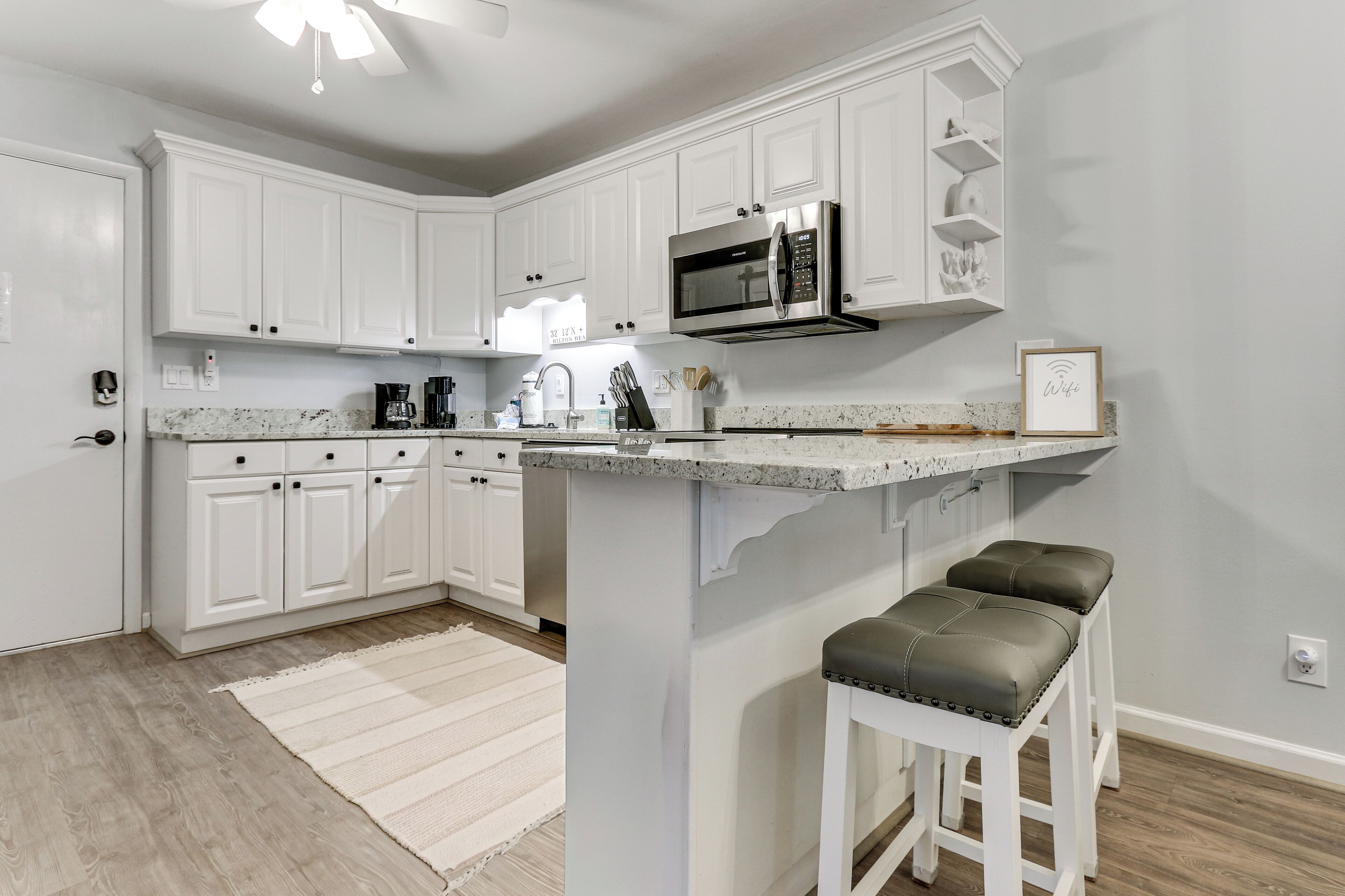A kitchen with white cabinets, a stainless steel microwave, a coffee maker, and a bar stool.