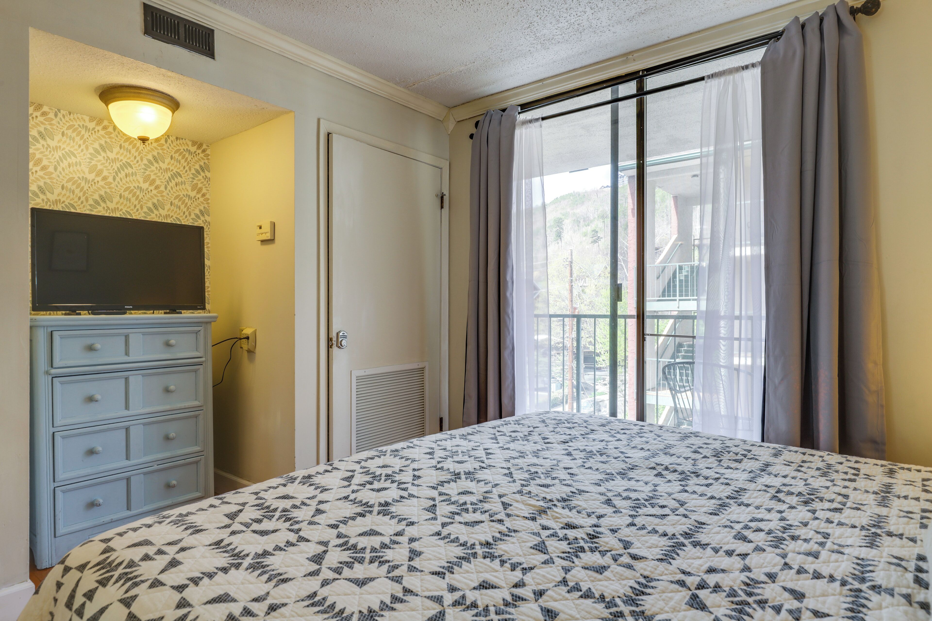 A bedroom with a bed, dresser, television, and a window with curtains.