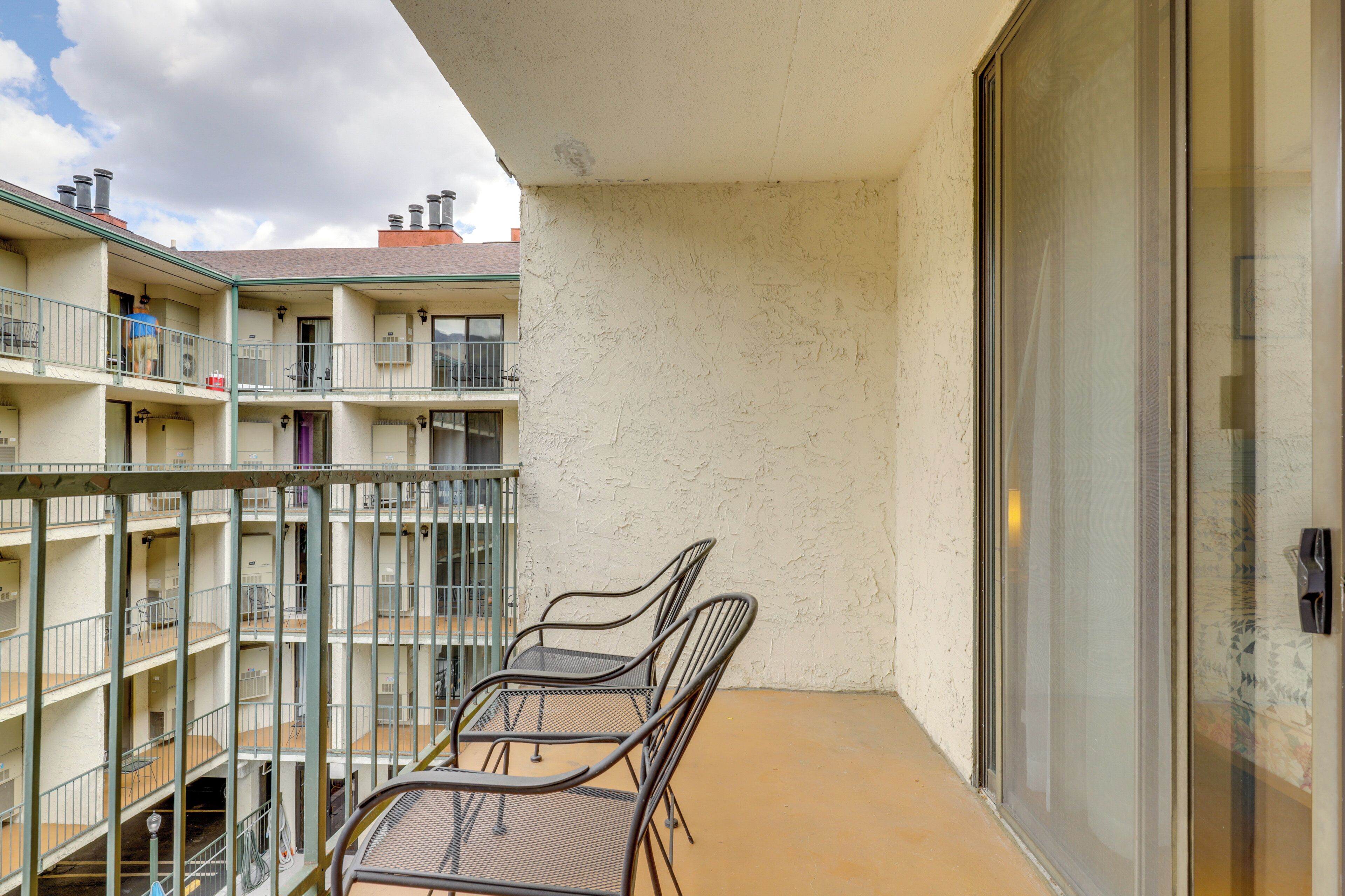 Un balcón con sillas y una mesa de metal, una puerta de cristal y vistas a un edificio de varios pisos con balcones.