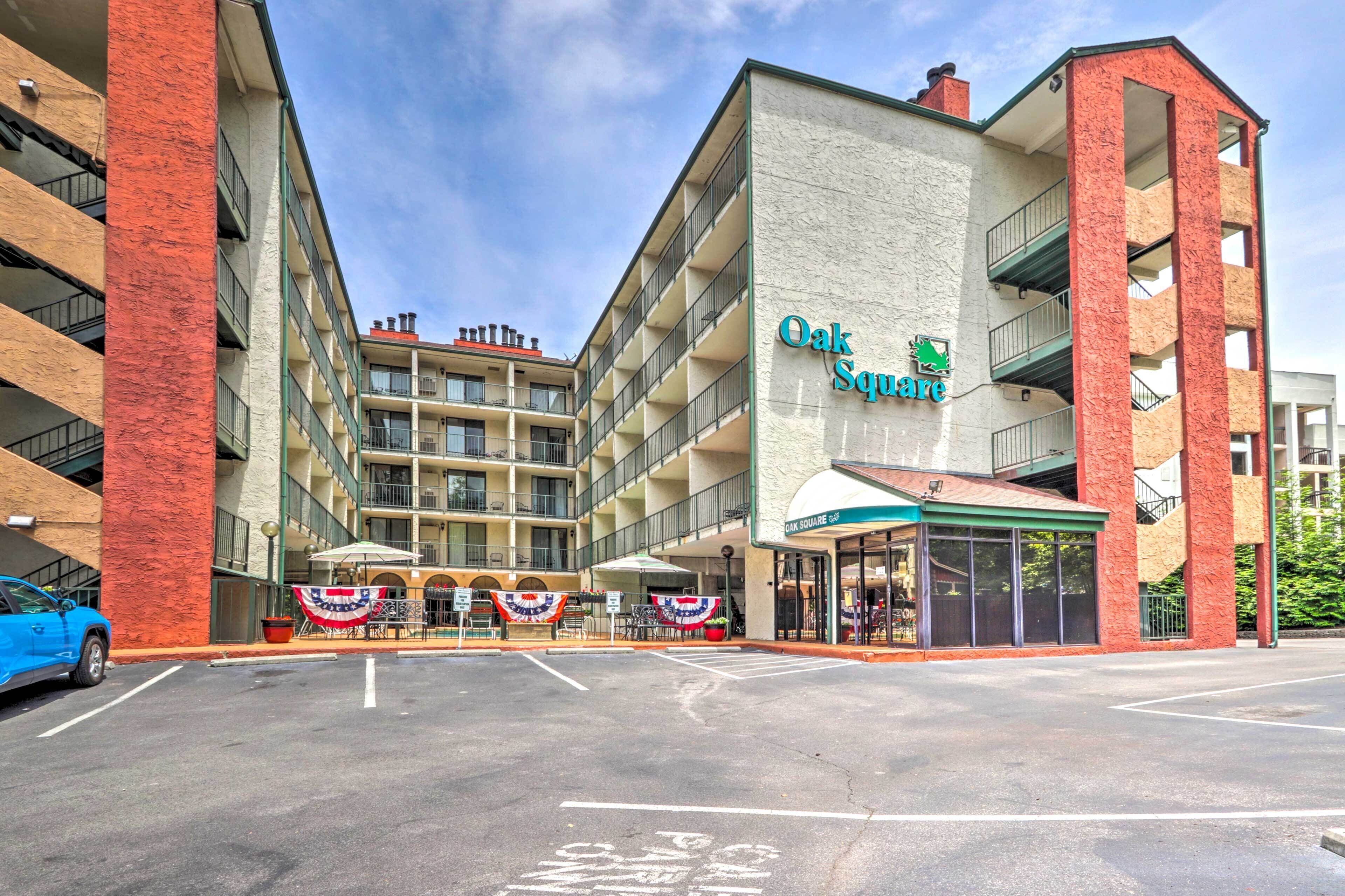 A multi-story hotel with a prominent sign reading 'Oak Square.' It features a parking lot, outdoor seating area with umbrellas, and a covered entrance.
