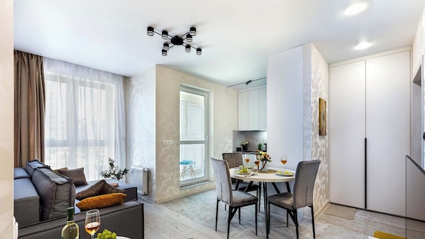 Een moderne woonkamer met een bank, een eettafel met stoelen, een keukenhoek en een groot raam met gordijnen.
