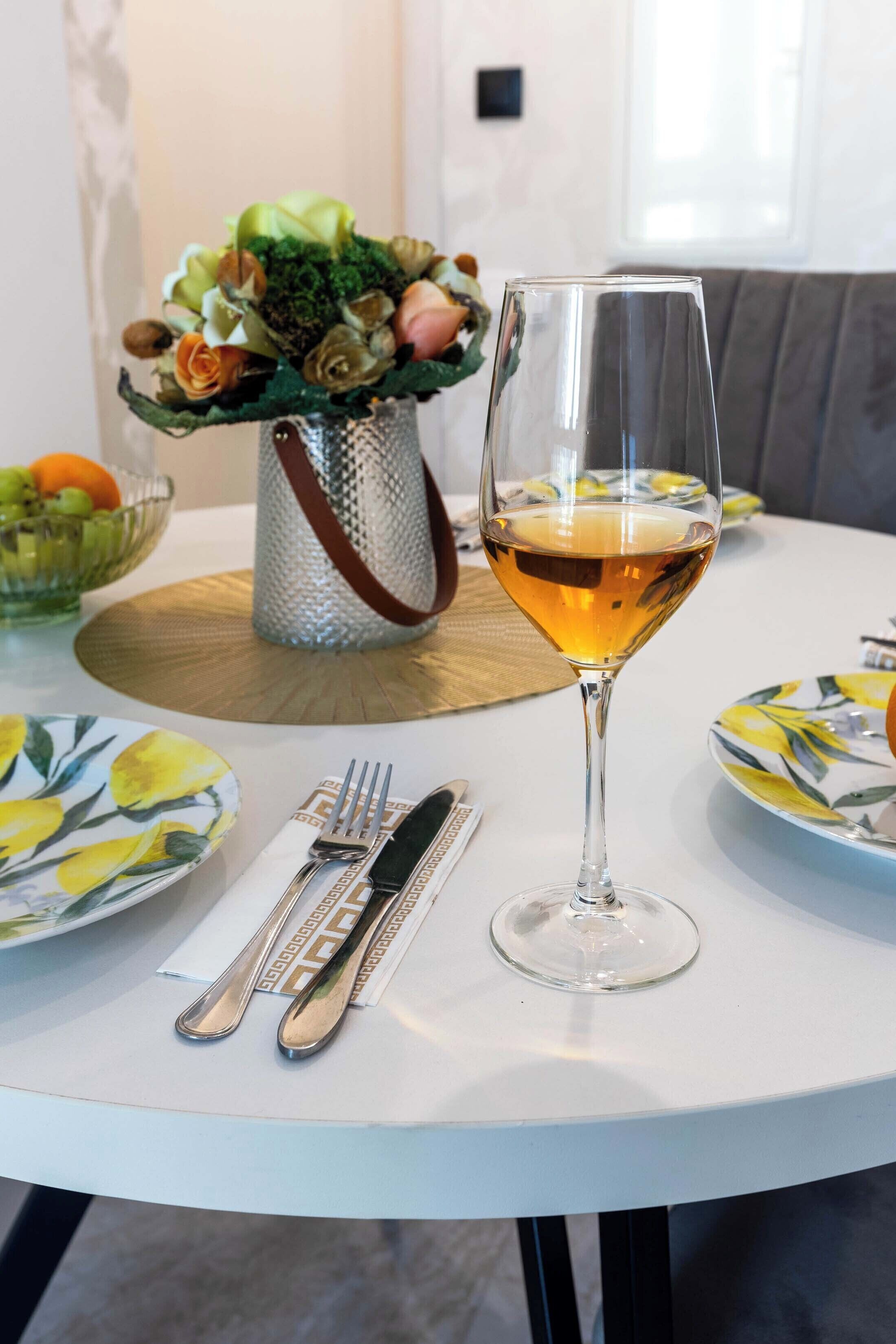 Una mesa de comedor con una copa de vino blanco, un jarrón con flores y un cuenco de fruta.