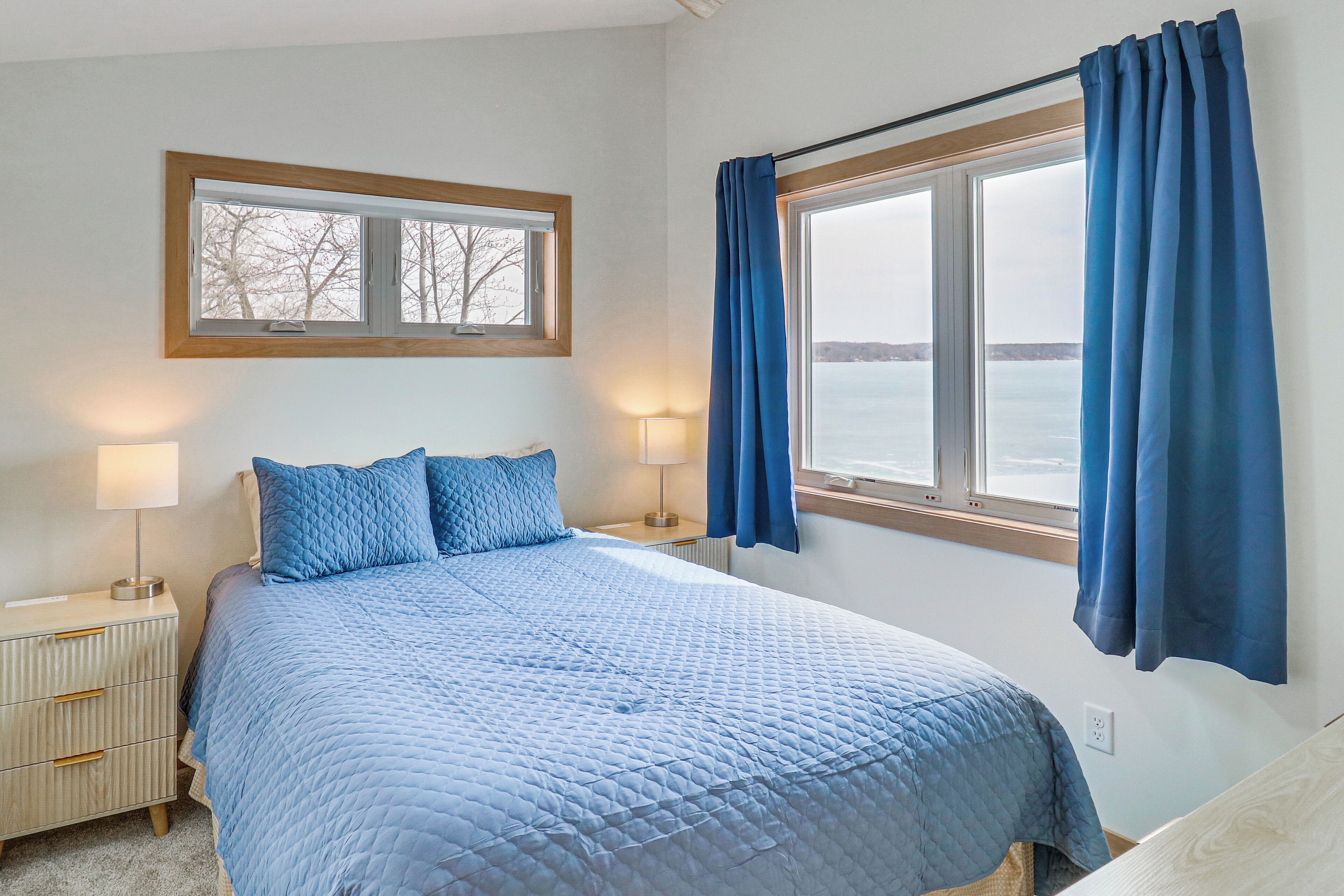 A bedroom with a bed, two bedside tables, a window with curtains, and a view of the water.