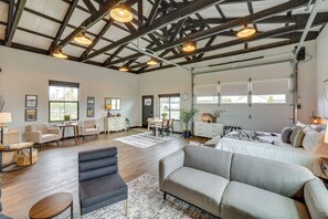 A spacious living area with a high ceiling, wooden beams, and large windows. There is a dining area with a table and chairs, a sofa, and a bed with a view of the garage. - Uncork & Unwind: Scenic Walla Walla Escape (Walla Walla)