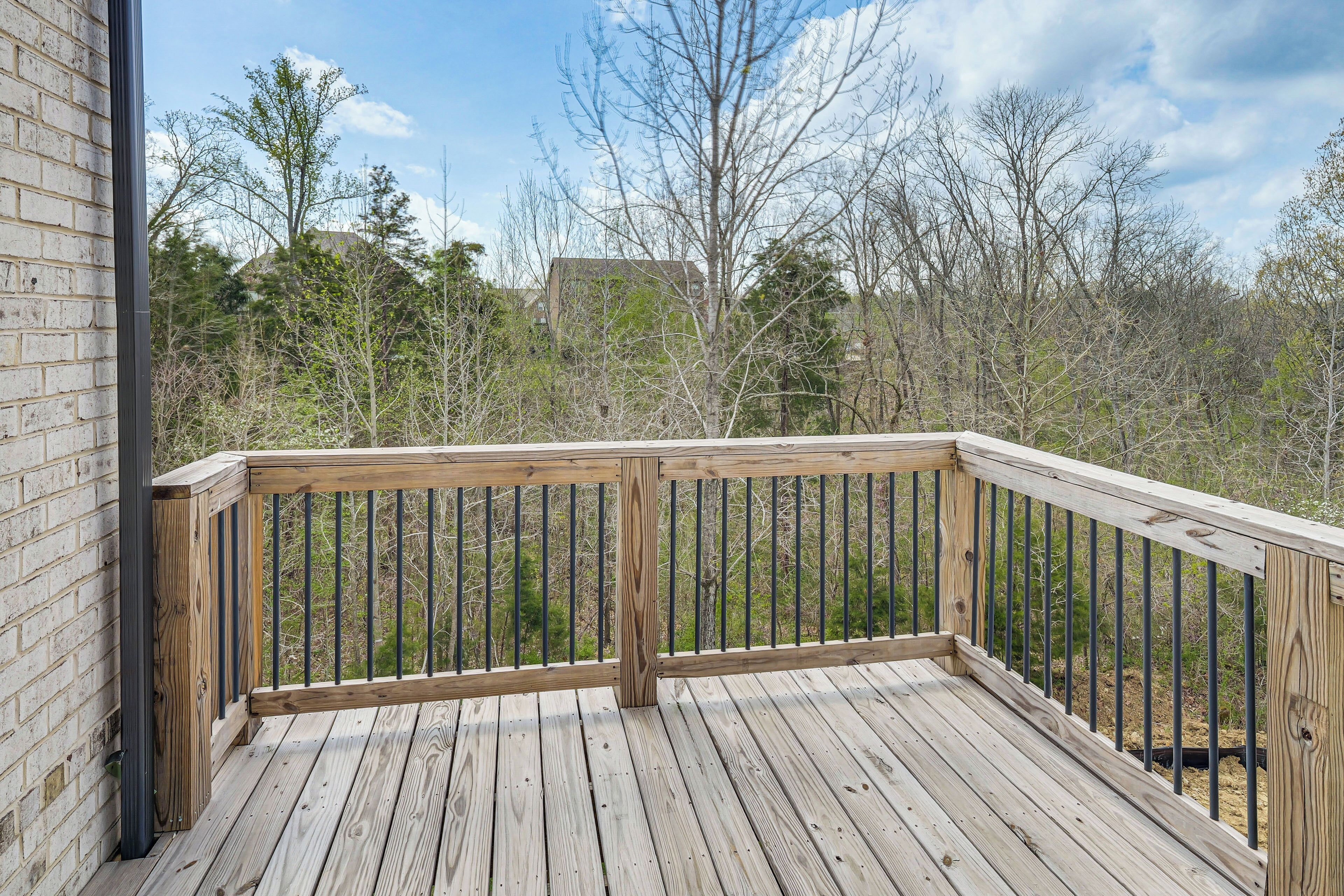 Une terrasse en bois avec une rambarde en métal, entourée d’arbres et avec un bâtiment visible au loin.