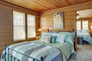 Quarto com painéis de madeira, uma cama grande, duas janelas, um quadro e uma mesa de cabeceira com abajur.