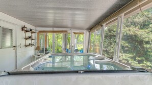 Ett bubbelbad med en glasvägg, en trähylla med diverse föremål och en utsikt över träd genom stora fönster.