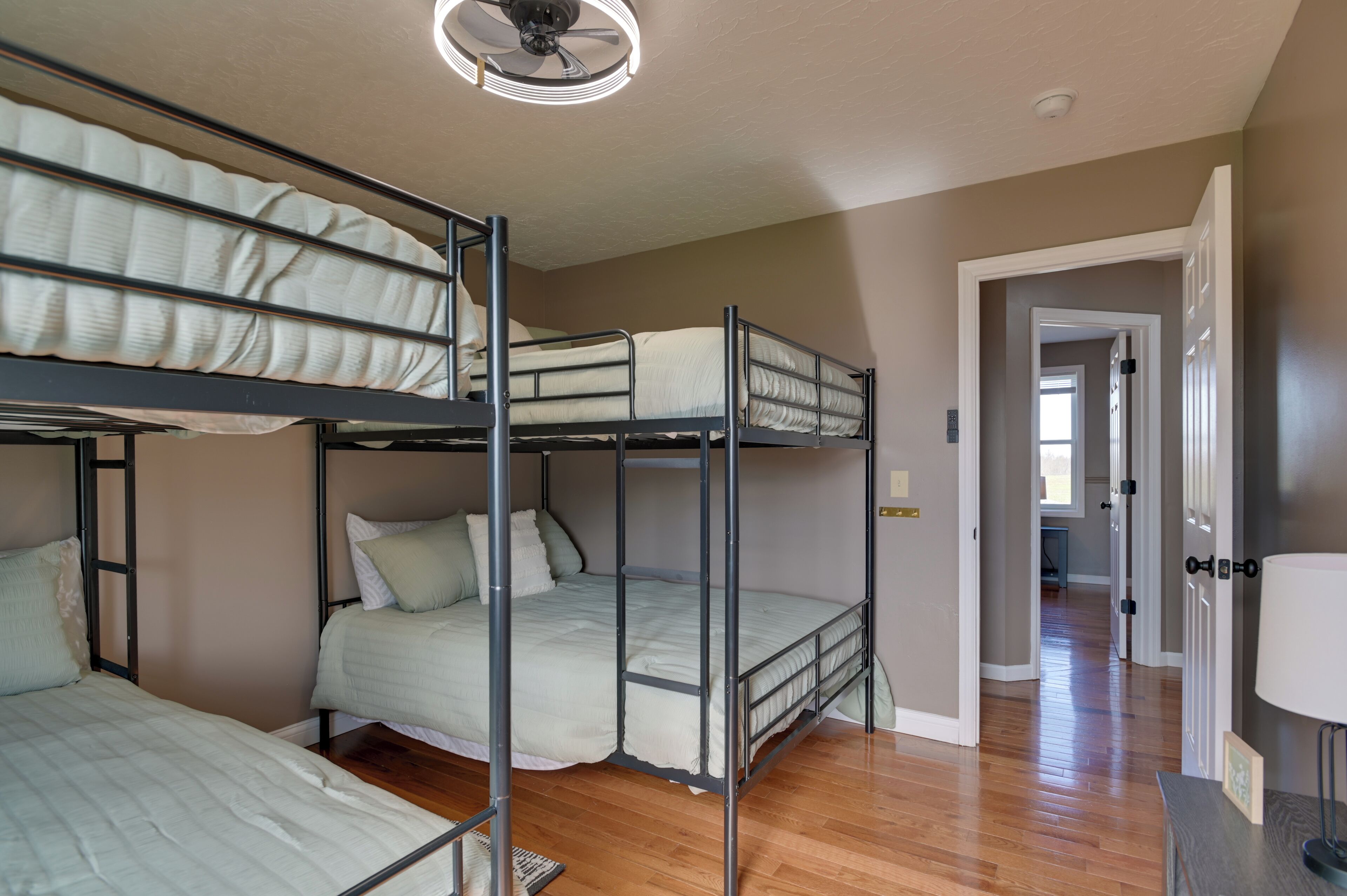 A bunk bed room with a ceiling fan, wooden flooring, and a hallway leading to another room.