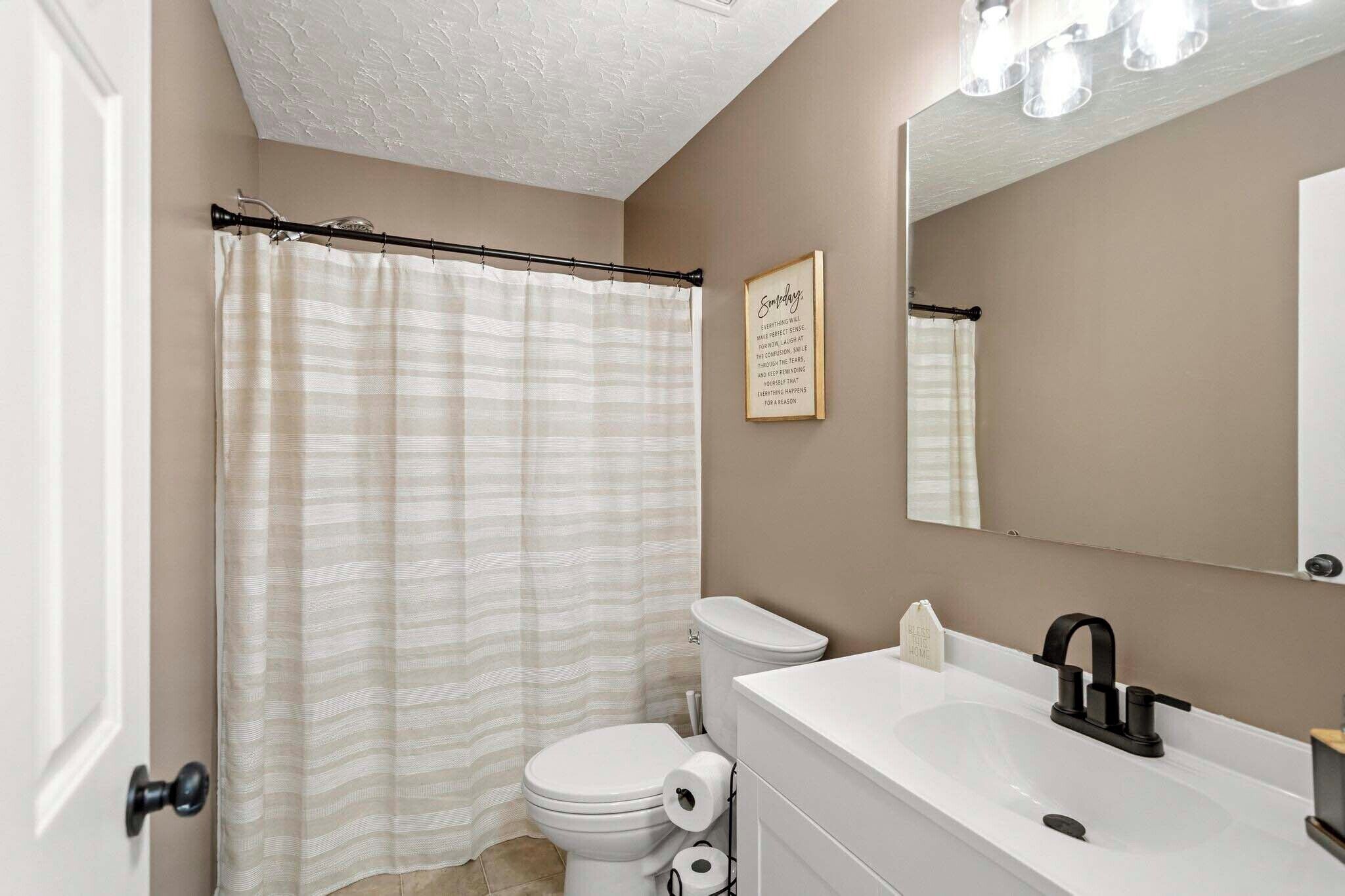 A bathroom with a striped shower curtain, a toilet, a sink with a black faucet, and a mirror with lights above it.