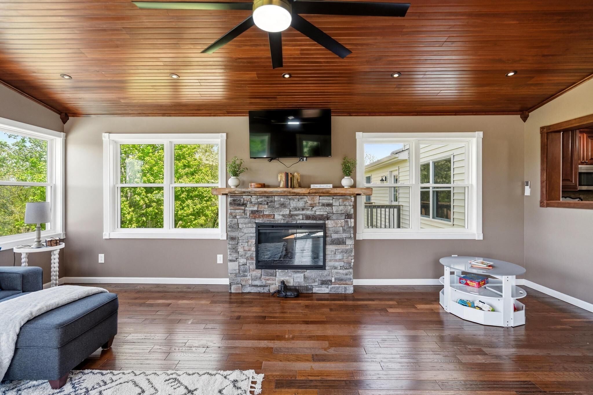 A living room with a stone fireplace, wooden ceiling, and large windows.